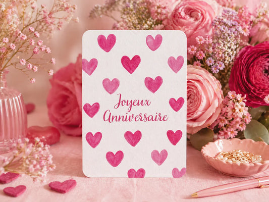 Carte Joyeux Anniversaire avec cœurs roses, entourée de fleurs et accessoires romantiques